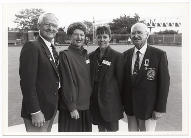 B&W photo of four people standing next to one another on a bowling green. In the background, there are a few people spread around on the green. In the distance, there are some large trees, a building, some fencing, and a bridge.
