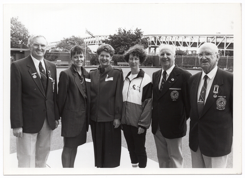 B&W photo of six people standing next to one another on a bowling green. In the distance, there are some large trees, a building, some fencing, and a bridge.