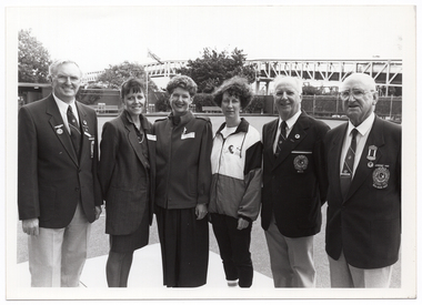 B&W photo of six people standing next to one another on a bowling green. In the distance, there are some large trees, a building, some fencing, and a bridge.
