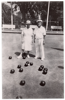 B&W photo of two people standing on a bowling green. They are wearing white uniforms, consisting of a white collared dress, a sunhat, and white shoes for the woman, and a white polo shirt, white trousers, a white cap, and white shoes for the man. There are several bowls (a ball for bowling) on the ground in front of them. Around the perimeter, there are three wooden benches on a low raised platform. Two shelters (partially obscured) are evident on the edges of the frame. There is a small scoreboard with a metal frame containing numbered cards that can be hung from the frame. There is a sign at the base of the frame, but the text and logos are illegible. In the background, there is a metal wire cyclone fence, with a few shrubs and small flowering plants in front of it and several large trees and shrubs behind it.