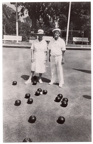 B&W photo of two people standing on a bowling green. They are wearing white uniforms, consisting of a white collared dress, a sunhat, and white shoes for the woman, and a white polo shirt, white trousers, a white cap, and white shoes for the man. There are several bowls (a ball for bowling) on the ground in front of them. Around the perimeter, there are three wooden benches on a low raised platform. Two shelters (partially obscured) are evident on the edges of the frame. There is a small scoreboard with a metal frame containing numbered cards that can be hung from the frame. There is a sign at the base of the frame, but the text and logos are illegible. In the background, there is a metal wire cyclone fence, with a few shrubs and small flowering plants in front of it and several large trees and shrubs behind it.