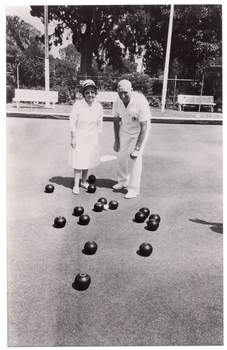 B&W photo of two people standing on a bowling green. They are wearing white uniforms, consisting of a white collared dress, a sunhat, and white shoes for the woman, and a white polo shirt, white trousers, and white shoes for the man. There are several bowls (a ball for bowling) on the ground in front of them. Around the perimeter, there are three wooden benches on a low raised platform. Two shelters (partially obscured) are evident on the edges of the frame. There is a small scoreboard (partially obscured) with a metal frame containing numbered cards that can be hung from the frame. In the background, there is a metal wire cyclone fence, with a few shrubs and small flowering plants in front of it and several large trees and shrubs behind it.