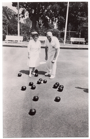 B&W photo of two people standing on a bowling green. They are wearing white uniforms, consisting of a white collared dress, a sunhat, and white shoes for the woman, and a white polo shirt, white trousers, and white shoes for the man. There are several bowls (a ball for bowling) on the ground in front of them. Around the perimeter, there are three wooden benches on a low raised platform. Two shelters (partially obscured) are evident on the edges of the frame. There is a small scoreboard (partially obscured) with a metal frame containing numbered cards that can be hung from the frame. In the background, there is a metal wire cyclone fence, with a few shrubs and small flowering plants in front of it and several large trees and shrubs behind it.