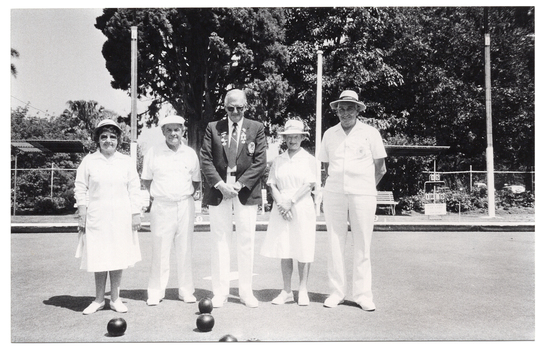 B&W photo of five people standing next to one another on a bowling green. In the centre, there is a man wearing a blazer jacket with medals and badges, a shirt and tie, white trousers, and white shoes. On either side of him, there is a man and a woman. The women are wearing white collared dresses, a sunhat, and white shoes. The men are wearing white polo shirts, white trousers, and white cap or sunhat, and white shoes. In the background, there are some wooden benches, two shelters, three tall wooden posts, a metal-framed scoreboard from which numbered cards can be hung, and several large trees behind a cyclone fence.