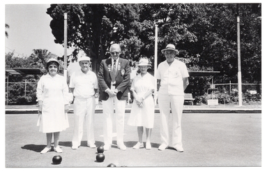 B&W photo of five people standing next to one another on a bowling green. In the centre, there is a man wearing a blazer jacket with medals and badges, a shirt and tie, white trousers, and white shoes. On either side of him, there is a man and a woman. The women are wearing white collared dresses, a sunhat, and white shoes. The men are wearing white polo shirts, white trousers, and white cap or sunhat, and white shoes. In the background, there are some wooden benches, two shelters, three tall wooden posts, a metal-framed scoreboard from which numbered cards can be hung, and several large trees behind a cyclone fence.