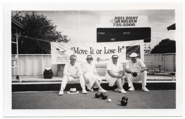 B&W photo of four people sitting on a ledge surrounding a bowling green. One person is holding a bowl, and there are several other bowls on the court in front of them. Behind them, there is a large "Move it or Lose it" horizontal banner with the text and logo for the Arthritis Foundation of Victoria and VicHealth Foundation at either end. Each end of the banner has been tied with rope to the poles of a covered shelter. There is a bench seat underneath each shelter. Behind the banner, there is a scoreboard with an advertising sign above it for "Noel Dight Holden". Below the advertising sign, the text 'LILYDALE', 'VISITORS', and 'ENDS' are evident on the scoreboard. In the background, there are a few large trees behind a fence.