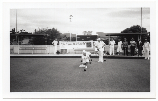 B&W photo of three people on the bowling green: a woman is bowling while two men look on, one of whom is holding a bowling ball in his hand. There is a group of people standing on a low raised platform on the edge of the green. Behind them, there is a large "Move it or Lose it" horizontal banner with the text and logo for the Arthritis Foundation of Victoria and VicHealth Foundation at either end. Each end of the banner has been tied with rope to the poles of a covered shelter. There is a bench seat underneath each shelter. Behind the banner, there is a scoreboard with an advertising sign above it for Noel Dight Holden. Below the advertising sign, the text 'LILYDALE', 'VISITORS', and 'ENDS' are evident on the scoreboard. There are a few trees and a house or building behind a fence, and some hills in the distance.