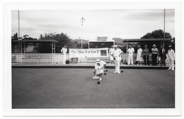 B&W photo of three people on the bowling green: a woman is bowling while two men look on, one of whom is holding a bowling ball in his hand. There is a group of people standing on a low raised platform on the edge of the green. Behind them, there is a large "Move it or Lose it" horizontal banner with the text and logo for the Arthritis Foundation of Victoria and VicHealth Foundation at either end. Each end of the banner has been tied with rope to the poles of a covered shelter. There is a bench seat underneath each shelter. Behind the banner, there is a scoreboard with an advertising sign above it for Noel Dight Holden. Below the advertising sign, the text 'LILYDALE', 'VISITORS', and 'ENDS' are evident on the scoreboard. There are a few trees and a house or building behind a fence, and some hills in the distance.