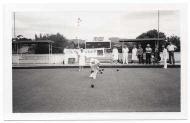 B&W photo of three people on the bowling green: a man is bowling while a woman and a man look on. There is a group of people standing on a low raised platform on the edge of the green. Behind them, there is a large "Move it or Lose it" horizontal banner with the text and logo for the Arthritis Foundation of Victoria and VicHealth Foundation at either end. Each end of the banner has been tied with rope to the poles of a covered shelter. There is a bench seat underneath each shelter. Behind the banner, there is a scoreboard with an advertising sign above it for Noel Dight Holden. Below the advertising sign, the text 'LILYDALE', 'VISITORS', and 'ENDS' are evident on the scoreboard. There are a few trees and a house or building behind a fence, and some hills in the distance.