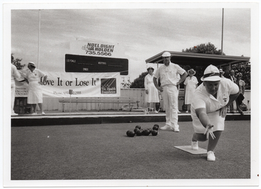 B&W photo of two people on the bowling green: a woman is bowling while a man, standing behind her, looks on. There is a group of people standing on a low raised platform on the edge of the green. Behind them, there is a large "Move it or Lose it" horizontal banner with the text and logo for the Arthritis Foundation of Victoria and VicHealth Foundation at either end. One end of the banner has been tied with rope to the poles of a covered shelter. There is a bench seat underneath the shelter (partially obscured). Behind the banner, there is a scoreboard with an advertising sign above it for Noel Dight Holden. Below the advertising sign, the text 'LILYDALE', 'VISITORS', and 'ENDS' are evident on the scoreboard. There are a few trees behind a fence.