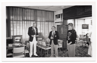 B&W photo of four people standing inside a clubhouse, (one person is on the edge of the frame, partially obscured). There are two club officials wearing club blazers and medals and a woman wearing a 'Move it or Lose it' sweatshirt. One of the club officials is looking down at a piece of paper that he is holding in his hands. There are several tables and chairs around the room. Around the clubhouse, there is a range of sports paraphanalia such as penant flags, an 'ACHIEVEMENTS' board, and a LADIES PENNANT scoreboard. In the background, there is a television set on a tall stand.