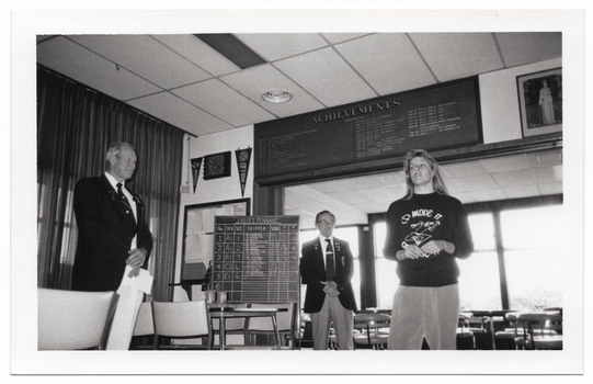 B&W photo of four people standing inside a clubhouse. There are two club officials wearing club blazers and medals and a woman wearing a 'Move it or Lose it' sweatshirt. The two club officials are looking at the woman. One of the club officials is holding a piece of paper in his hands. There is a range of sports paraphanalia such as penant flags, an 'ACHIEVEMENTS' board, a full length portrait of Queen Elizabeth II, a LADIES PENNANT scoreboard, and several tables and chairs around the room. This photo has been taken from a low angle.