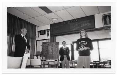 B&W photo of four people standing inside a clubhouse. There are two club officials wearing club blazers and medals and a woman wearing a 'Move it or Lose it' sweatshirt. The two club officials are looking at the woman. One of the club officials is holding a piece of paper in his hands. There is a range of sports paraphanalia such as penant flags, an 'ACHIEVEMENTS' board, a full length portrait of Queen Elizabeth II, a LADIES PENNANT scoreboard, and several tables and chairs around the room. This photo has been taken from a low angle.