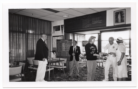 B&W photo of five people standing inside a clubhouse. There are two club officials wearing club blazers and medals, a woman wearing a 'Move it or Lose it' sweatshirt, and two people wearing white lawn bowls uniforms. The woman is handing a piece of paper to the two lawn bowls players. There is a range of sports paraphanalia such as penant flags, an 'ACHIEVEMENTS' board, a full length portrait of Queen Elizabeth II, a LADIES PENNANT scoreboard, and several tables and chairs around the room. In the background, there is a television set on a tall stand.