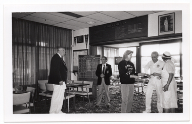 B&W photo of five people standing inside a clubhouse. There are two club officials wearing club blazers and medals, a woman wearing a 'Move it or Lose it' sweatshirt, and two people wearing white lawn bowls uniforms. The woman is handing a piece of paper to the two lawn bowls players. There is a range of sports paraphanalia such as penant flags, an 'ACHIEVEMENTS' board, a full length portrait of Queen Elizabeth II, a LADIES PENNANT scoreboard, and several tables and chairs around the room. In the background, there is a television set on a tall stand.