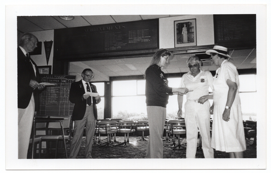 B&W photo of five people standing inside a clubhouse. There are two club officials wearing club blazers and medals, a woman wearing a 'Move it or Lose it' sweatshirt, and two people wearing white lawn bowls uniforms. The woman is handing a piece of paper to the two bowls players. The two club officials are each looking down at a piece of paper in their hands. There is a range of sports paraphanalia such as penant flags, an 'ACHIEVEMENTS' board, a full length portrait of Queen Elizabeth II, a LADIES PENNANT scoreboard, and several tables and chairs around the room.