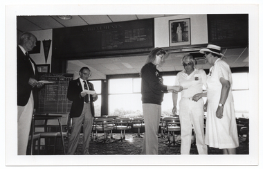 B&W photo of five people standing inside a clubhouse. There are two club officials wearing club blazers and medals, a woman wearing a 'Move it or Lose it' sweatshirt, and two people wearing white lawn bowls uniforms. The woman is handing a piece of paper to the two bowls players. The two club officials are each looking down at a piece of paper in their hands. There is a range of sports paraphanalia such as penant flags, an 'ACHIEVEMENTS' board, a full length portrait of Queen Elizabeth II, a LADIES PENNANT scoreboard, and several tables and chairs around the room.