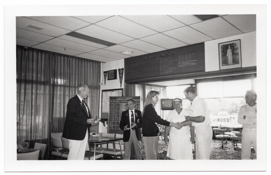 B&W photo of six people standing inside a clubhouse. There are two club officials wearing club blazers and medals, a woman wearing a 'Move it or Lose it' sweatshirt, and three people wearing white lawn bowls uniforms. The woman is shaking hands with a lawn bowls player while the others look on. There is a range of sports paraphanalia such as penant flags, an 'ACHIEVEMENTS' board, a full length portrait of Queen Elizabeth II, a LADIES PENNANT scoreboard, and several tables and chairs around the room. In the background, there is a television set on a tall stand.