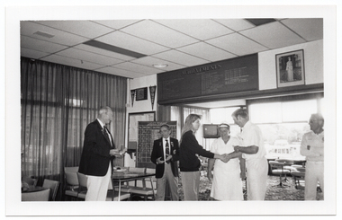 B&W photo of six people standing inside a clubhouse. There are two club officials wearing club blazers and medals, a woman wearing a 'Move it or Lose it' sweatshirt, and three people wearing white lawn bowls uniforms. The woman is shaking hands with a lawn bowls player while the others look on. There is a range of sports paraphanalia such as penant flags, an 'ACHIEVEMENTS' board, a full length portrait of Queen Elizabeth II, a LADIES PENNANT scoreboard, and several tables and chairs around the room. In the background, there is a television set on a tall stand.