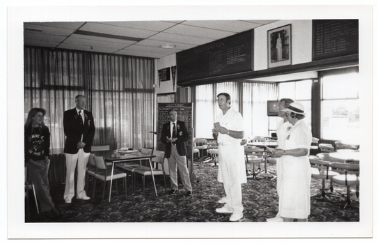 B&W photo of seven people standing inside a clubhouse (one partially obscured). There are two club officials wearing club blazers and medals, a woman wearing a 'Move it or Lose it' sweatshirt, and four people wearing white lawn bowls uniforms. One of the bowls players is speaking while the others look on. There is a range of sports paraphanalia such as penant flags, an 'ACHIEVEMENTS' board, a full length portrait of Queen Elizabeth II, a LADIES PENNANT scoreboard, and several tables and chairs around the room. In the background, there is a television set on a tall stand.
