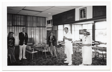 B&W photo of seven people standing inside a clubhouse (one partially obscured). There are two club officials wearing club blazers and medals, a woman wearing a 'Move it or Lose it' sweatshirt, and four people wearing white lawn bowls uniforms. One of the bowls players is speaking while the others look on. There is a range of sports paraphanalia such as penant flags, an 'ACHIEVEMENTS' board, a full length portrait of Queen Elizabeth II, a LADIES PENNANT scoreboard, and several tables and chairs around the room. In the background, there is a television set on a tall stand.