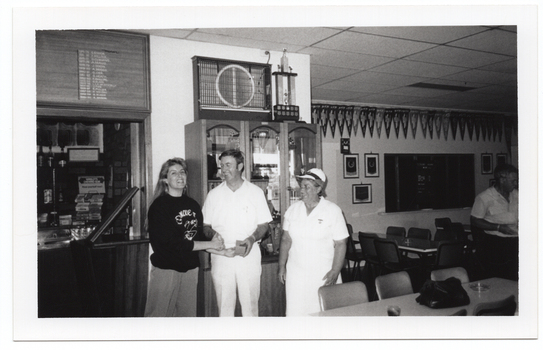 B&W photo of three people standing close to one another inside a clubhouse, with a fourth person visible near the edge of the frame. One is a woman wearing a 'Move it or Lose it' sweatshirt, and the other two people are wearing white lawn bowls uniforms. The fourth person (on the edge of the frame) is wearing a white shirt with dark coloured trousers. The woman is shaking hands with a man while he hands her a small box, which she takes with her other hand. There is a range of sports paraphanalia such as a trophy cabinet, penant flags and small framed banners on the walls, and several tables and chairs around the room.