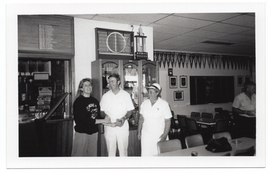 B&W photo of three people standing close to one another inside a clubhouse, with a fourth person visible near the edge of the frame. One is a woman wearing a 'Move it or Lose it' sweatshirt, and the other two people are wearing white lawn bowls uniforms. The fourth person (on the edge of the frame) is wearing a white shirt with dark coloured trousers. The woman is shaking hands with a man while he hands her a small box, which she takes with her other hand. There is a range of sports paraphanalia such as a trophy cabinet, penant flags and small framed banners on the walls, and several tables and chairs around the room.