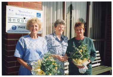 COL medium close up photo of three women standing in front of a red brick building. Two of the women are each holding a floral bouquet of yellow roses, miniature daisies, and green foliage, while the woman in the middle is holding a pair of spectacles. There is a small sign on the building behind them. It contains the text, "Move it or Lose it" Bowls helps beat Arthritis. On either side of this text is the text and logo for the Arthritis Foundation of Victoria and the Victorian Health Promotion Foundation (VicHealth). Following that, there is a succession of numbers, beginning with "10 11 12 13 14...", with the remainder partially obscured. Then, there is a logo for the Royal Victorian Bowls Association, the text "PACE", and the text "Another project funded by Victorian Health Promotion with [partially obscured] of Victoria". Behind the women, there is a pale green wooden bench beneath some large curtained windows.