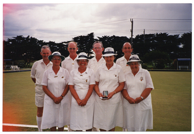 COL photo of a small group of eight people standing in two rows of four on a bowling green. One woman in the front row is holding a gold watch in a presentation box and a small piece of paper. In the background, shelters and wooden benches are evident around the perimeter of the grass court. There are several large trees in a row behind the bowling green. There is some orange coloured discolouration around the left edge of the frame on the image layer, due to early exposure to light of the undeveloped film.