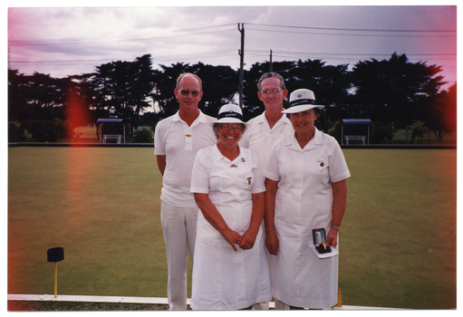 COL photo of a small group of four people standing in two rows of two on a bowling green. One woman in the front row is holding a gold watch in a presentation box and a small piece of paper. In the background, shelters and wooden benches can be seen around the perimeter of the grass court. There are several large trees in a row behind the bowling green. There is some orange coloured discolouration on the image layer, due to early exposure to light of the undeveloped film.