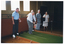 COL photo of a small group of people gathered around a portable bowling green (or practice rink). They are inside a room with polished floorboards, large windows, and a decorative dado feature on the painted walls. One man stands on the practice lawn, preparing to bowl, as the others look on. Behind him, there is a man standing, holding a small white ball. There are several bowling bowls at his feet on the practice green. Beside the pratice green, there is a queue rope barrier on some metal stands.