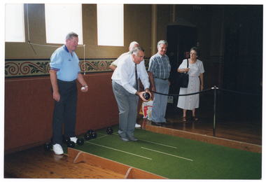 COL photo of a small group of people gathered around a portable bowling green (or practice rink). They are inside a room with polished floorboards, large windows, and a decorative dado feature on the painted walls. One man stands on the practice lawn, preparing to bowl, as the others look on. Behind him, there is a man standing, holding a small white ball. There are several bowling bowls at his feet on the practice green. Beside the pratice green, there is a queue rope barrier on some metal stands.
