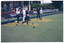 COL photo of a group of people at a lawn bowls club. One man is bowling while several men are standing on the green behind him. A few others are standing behind the green, beneath the veranda of a building and a woman is coming down the steps from the veranda towards the green. There is a small four-post frame near the edge of the court. A wooden sign affixed to a wooden retaining wall between the building and the grassy court contains the text "ROBERT W SMITH BOWLING GREEN".