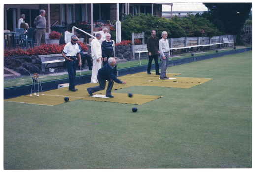 COL photo of a group of people at a lawn bowls club. One man is bowling while several men are standing on the green behind him. A few others are standing behind the green, beneath the veranda of a building and a woman is coming down the steps from the veranda towards the green. There is a small four-post frame near the edge of the court. A wooden sign affixed to a wooden retaining wall between the building and the grassy court contains the text "ROBERT W SMITH BOWLING GREEN".