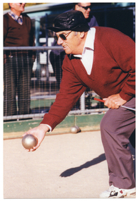 COL photo of a man, who is holding a silver bocce ball in his hand, crouching forward with one knee bent to bowl a silver ball. He is playing on a clay court. In the background, there are two men standing behind a wire fence. There are one or two silver balls against a wooden board at the edge of the court.