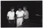 B&W photo of three women standing in behind a table tennis table. Two of the women are each holding a racquet or bat (also known as a 'paddle' in the USA) in one hand, while the third woman's hand is guiding the hand (and paddle) of one of the women.