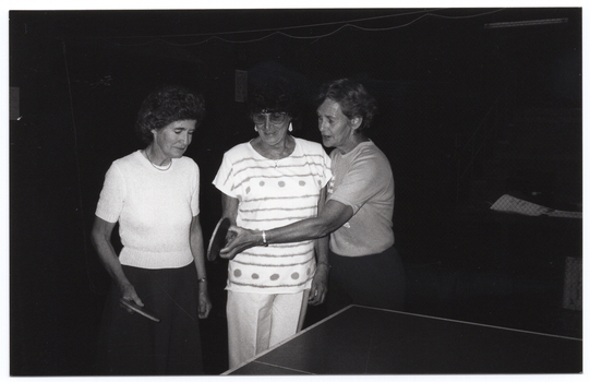 B&W photo of three women standing in behind a table tennis table. Two of the women are each holding a racquet or bat (also known as a 'paddle' in the USA) in one hand, while the third woman's hand is guiding the hand (and paddle) of one of the women.