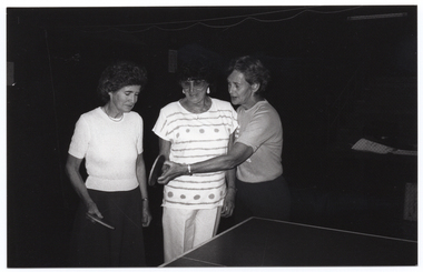B&W photo of three women standing in behind a table tennis table. Two of the women are each holding a racquet or bat (also known as a 'paddle' in the USA) in one hand, while the third woman's hand is guiding the hand (and paddle) of one of the women.