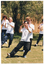 COL photo of a group of people, led by an instructor, practising Tai Chi on a grassy area. They are holding their hands up in front of their chests and leaning to one side with their weight on one leg, while stretching the other leg out to the other side. In the background, there is a wooden fence with a large tree behind it.