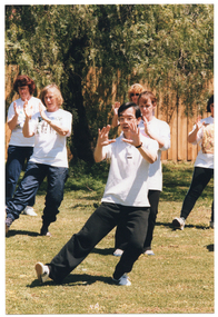 COL photo of a group of people, led by an instructor, practising Tai Chi on a grassy area. They are holding their hands up in front of their chests and leaning to one side with their weight on one leg, while stretching the other leg out to the other side. In the background, there is a wooden fence with a large tree behind it.