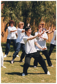 COL photo of a group of people, led by an instructor, practising Tai Chi on a grassy area. They are holding one hand outstretched in front of their chests and the other hand down nar the hip, while leaning to one side with their weight on one leg. In the background, there is a wooden fence with a large tree behind it.