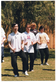 COL photo of a group of people, led by an instructor, practising Tai Chi on a grassy area. They are holding their hands down in front of their hips, with their fingers apart. The instructor is standing on one leg, with his other leg only slightly lifted off the ground. The three the class participants depicted still have both feet on the ground. In the background, there is a wooden fence with a large tree behind it.