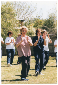 COL photo of a group of people practising Tai Chi on a grassy area. They are holding their hands up in front of their chests while standing on one leg, with the other leg lifted only slightly off the ground. In the background is a wooden fence with a large tree and a two-storey house behind it.