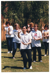 COL photo of a group of people, led by an instructor, practising Tai Chi on a grassy area. They are standing in rows in a triangle formation, with the instructor at the head. With both feet on the ground, they are holding one hand up (palm outward) in front of their faces, and one hand down (palm downward) in front of their stomachs. In the background, there is a wooden fence with a large tree and a brick building (mostly obscured) behind it.
