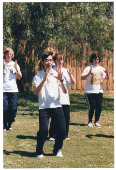 COL photo of a group of people, led by an instructor, practising Tai Chi on a grassy area. They are standing with both feet on the ground, knees slightly bent, while holding their hands up (palms facing) in front of their upper chests. In the background, there is a wooden fence with a large tree behind it.