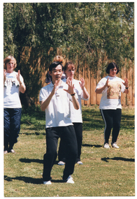 COL photo of a group of people, led by an instructor, practising Tai Chi on a grassy area. They are standing with both feet on the ground, knees slightly bent, while holding their hands up (palms facing) in front of their upper chests. In the background, there is a wooden fence with a large tree behind it.