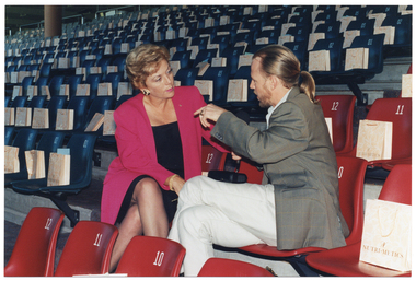 COL photo of two people sitting on two seats in rows of empty tiered stadium seating. They are facing one another, and the man is talking to the woman. He is wearing a grey checked blazer over white shirt and trousers and has his long hair tied back in a ponytail. She is wearing a bright pink blazer over a black dress. There is a Nutrimetics gift bag on every empty seat.