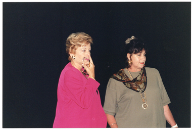 COL medium close up photo of two women, standing close to one another. One woman is wearing a bright pink blazer jacket. She is holding her spectacles in her hand while touching her nose with her finger. The other woman is wearing an olive green top with a brown and black scarf and some long necklaces. The background is completely black.
