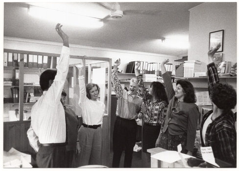 B&W photo of a small group of people doing some stretching exercises. Standing in a semi-circle, they are extending one arm out straight up towards the ceiling. They are in an office setting, with multiple shelves of folders, publications, and other office-related items behind them.