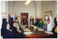 COL photo of a group of people, seated around a long wooden table. They are stretching their arms above their heads with their fingers interlocked and palms turned upwards. Each person has some paperwork on the table in front of them. There are also some pens, a few mugs, a few pairs of spectacles, and a polystyrene cup on the table.